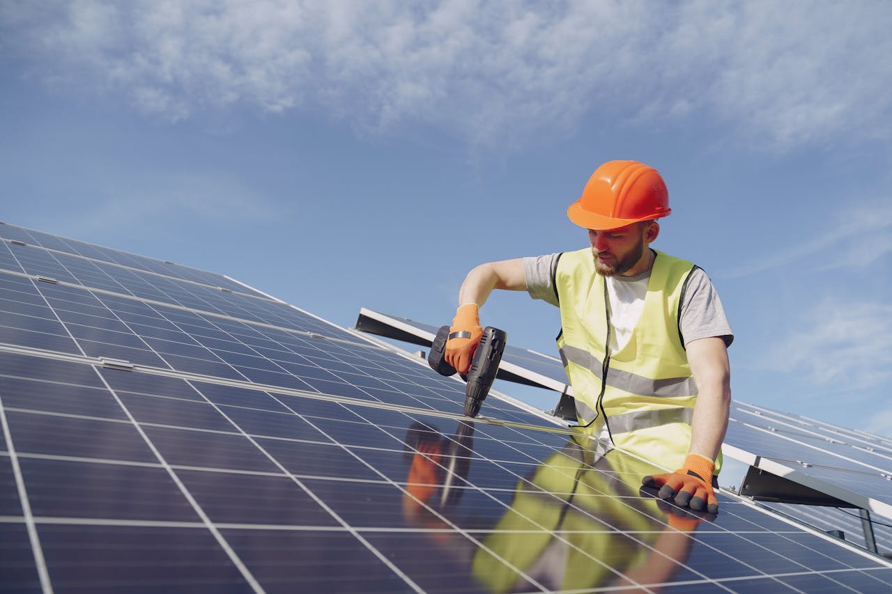 From below of concentrated young bearded male technician in protective clothes mounting solar batteries using drill against blue sky
